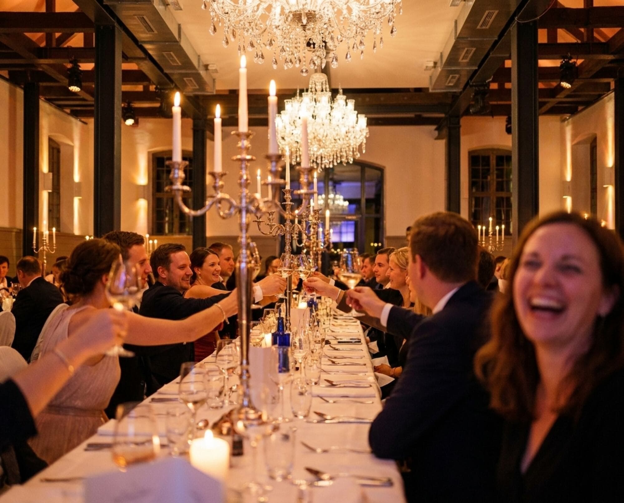 Festlich gekleidete Gäste stoßen an einer langen, weiß eingedeckten Tafel miteinander an, darüber die Kronleuchter und das historische Holzgebälk im warmen Kerzenlicht