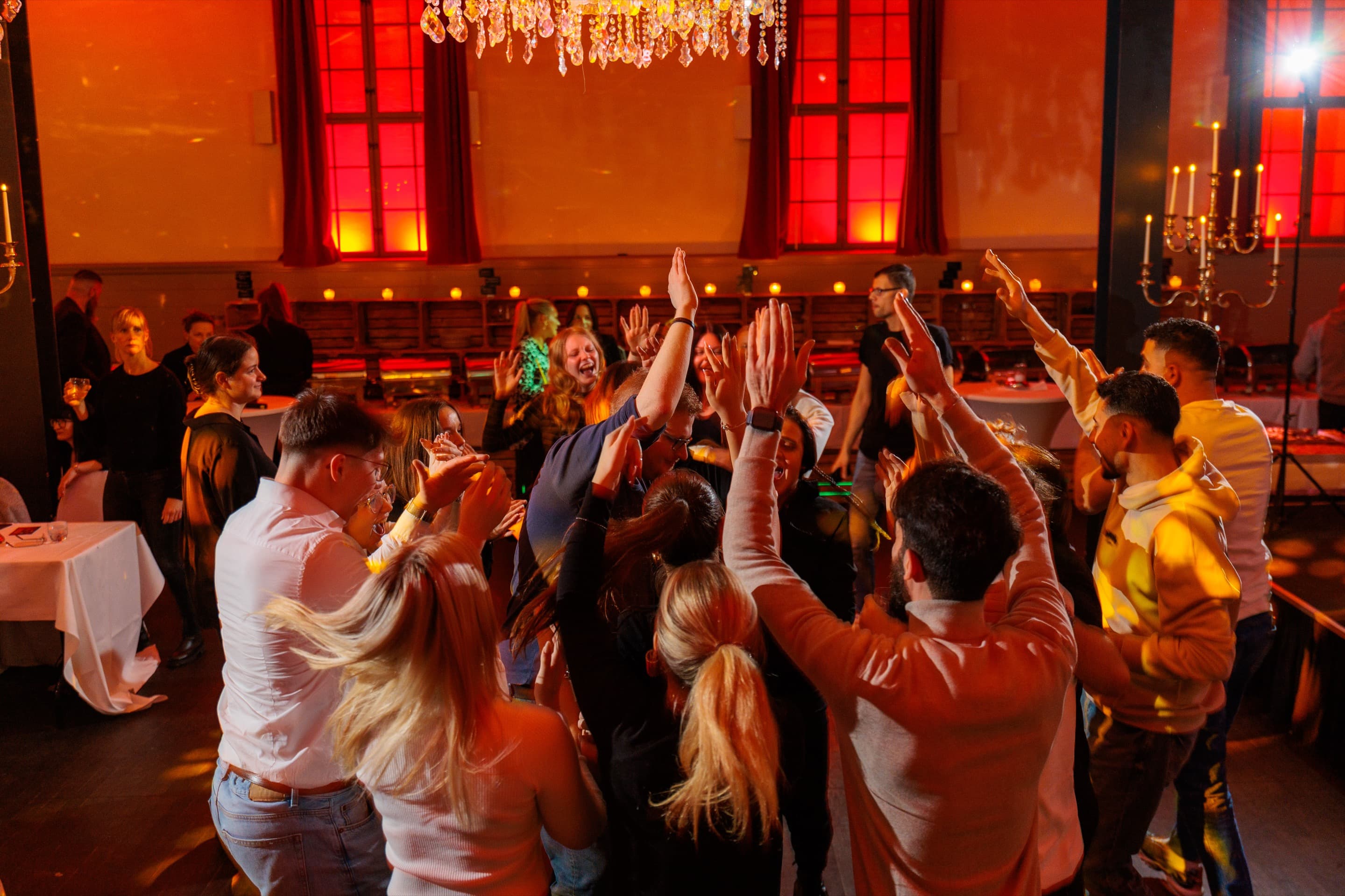Volle Tanzfläche im Saal, junge Gäste mit erhobenen Armen unter dem Kronleuchter, die hohen Fenster rot angestrahlt
