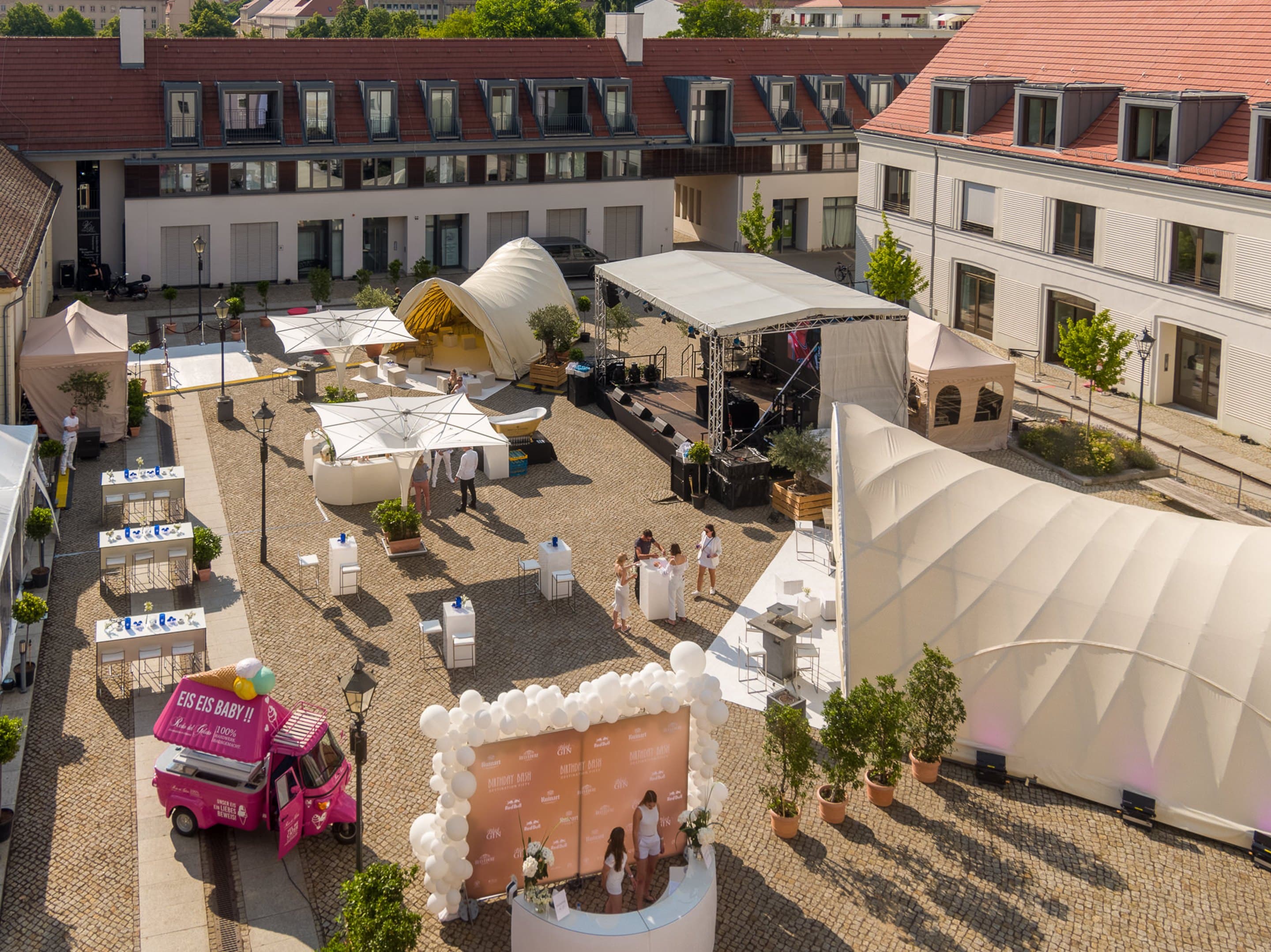 Derselbe Innenhof bei Tageslicht, Bühne, weiße Zelte und Sonnenschirme, Stehtische und der pinke Eiswagen auf dem Kopfsteinpflaster