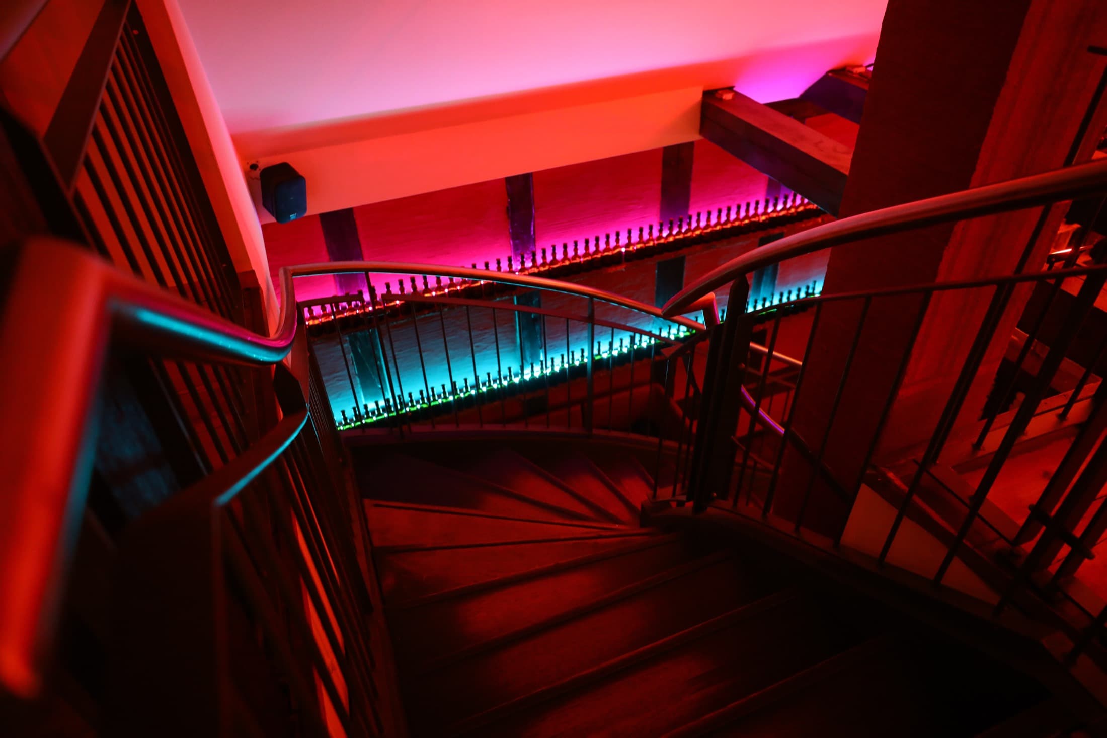 Blick von oben in eine gewendelte Holztreppe, der Handlauf von unten türkis beleuchtet, die Wände in Rot