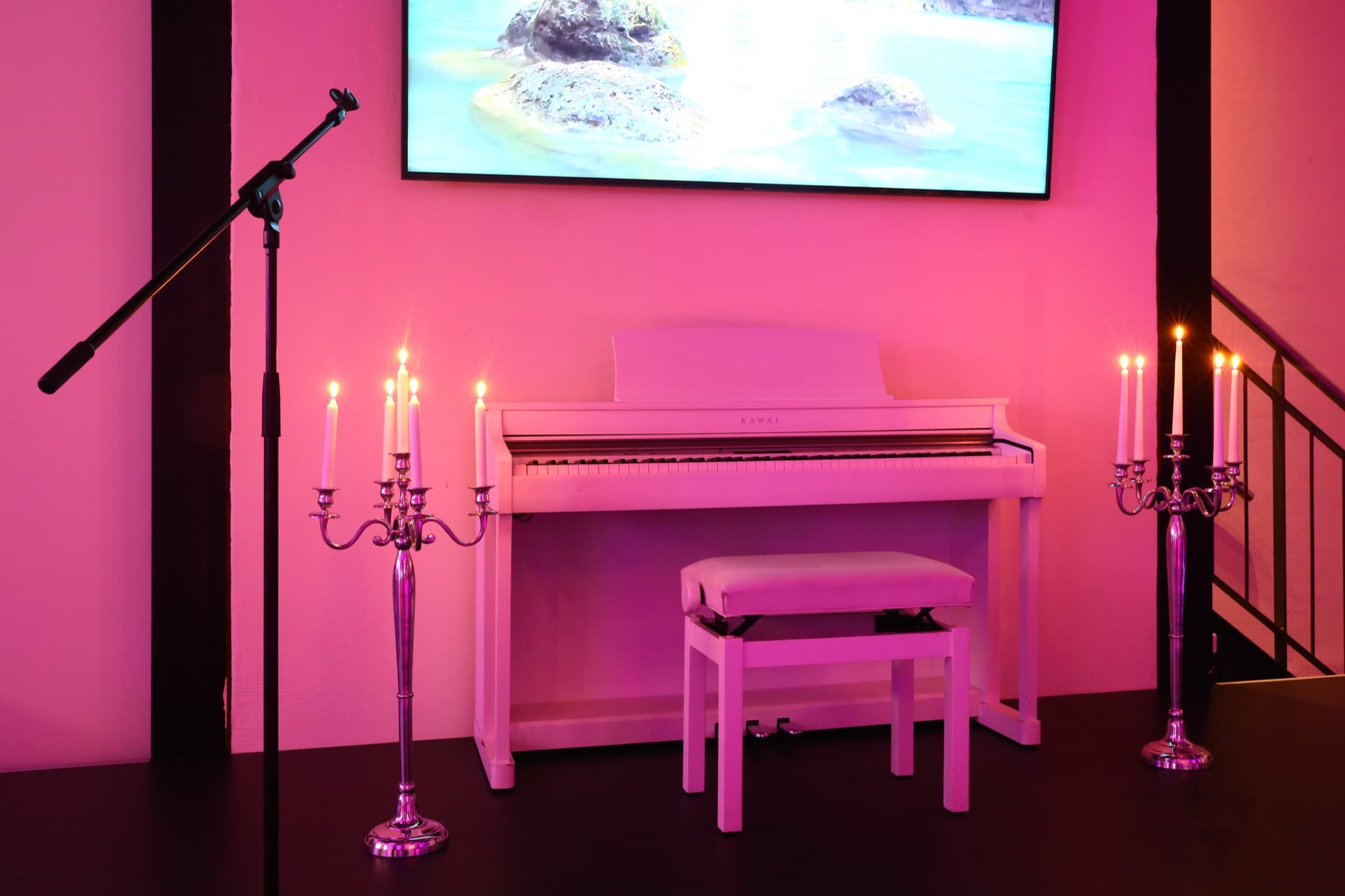 Ein weißes Piano mit Hocker vor pink beleuchteter Wand, daneben ein silberner Kerzenleuchter mit brennenden Kerzen