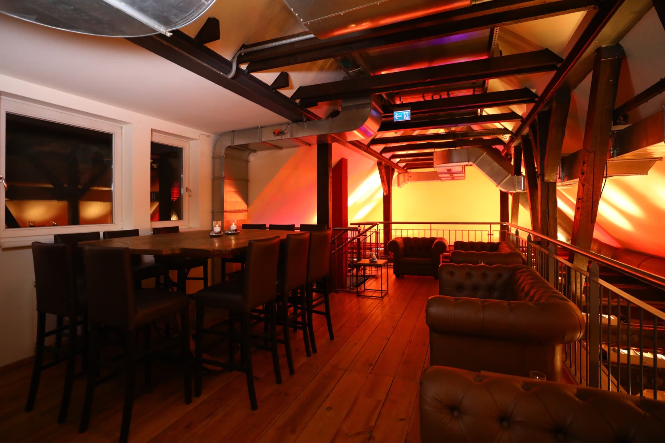 Das Obergeschoss im Hofmeister, links eine lange Tafel mit Barhockern, rechts Ledersofas an der Brüstung, das Gebälk orange angestrahlt