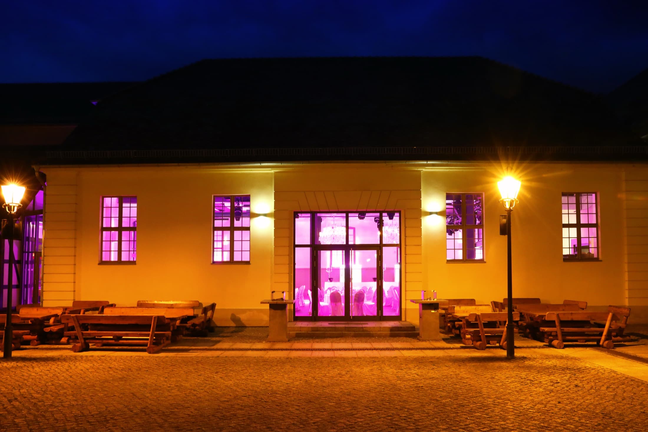 Die Fassade bei Nacht, die hohen Fenster von innen pink erleuchtet, davor Bänke auf dem Kopfsteinpflaster und eine Laterne