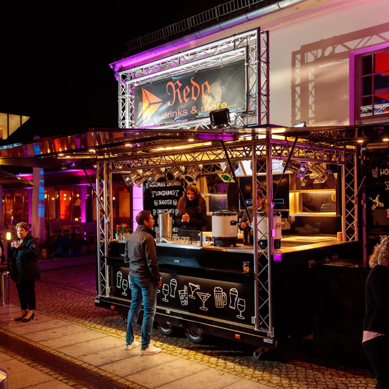Der Getränkestand Redo Drinks and More bei Nacht, ein Gerüst mit Lichtern über dem Tresen, dahinter eine Mitarbeiterin, davor ein Gast