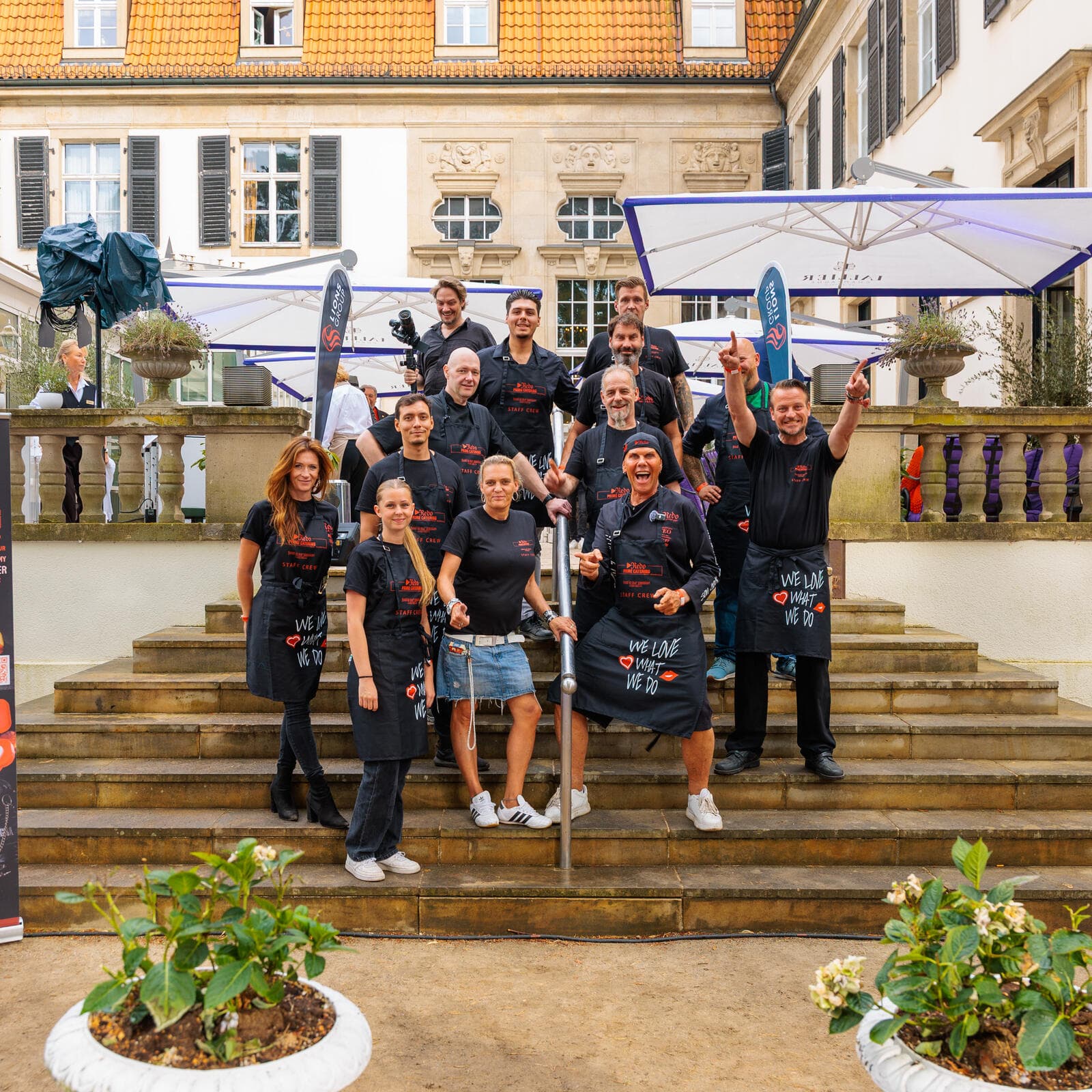 Das Team in schwarzen Redo-Schürzen auf einer Freitreppe, daneben das Banner von Redo Prime Catering