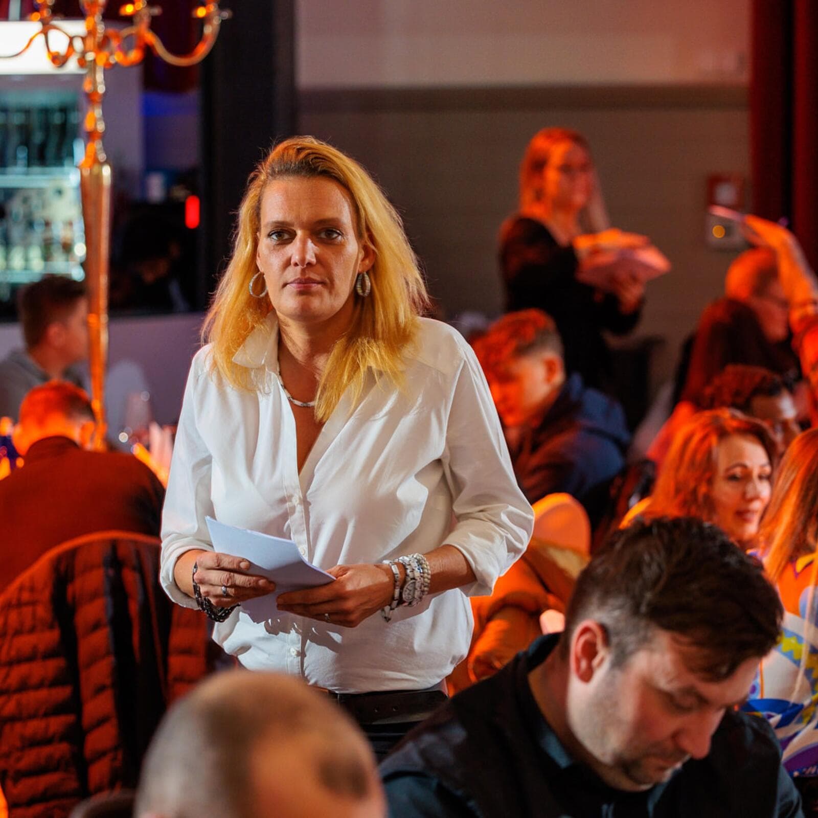Katharina Dögnitz in weißer Bluse mit Notizen in der Hand, konzentriert zwischen den Tischen einer laufenden Veranstaltung