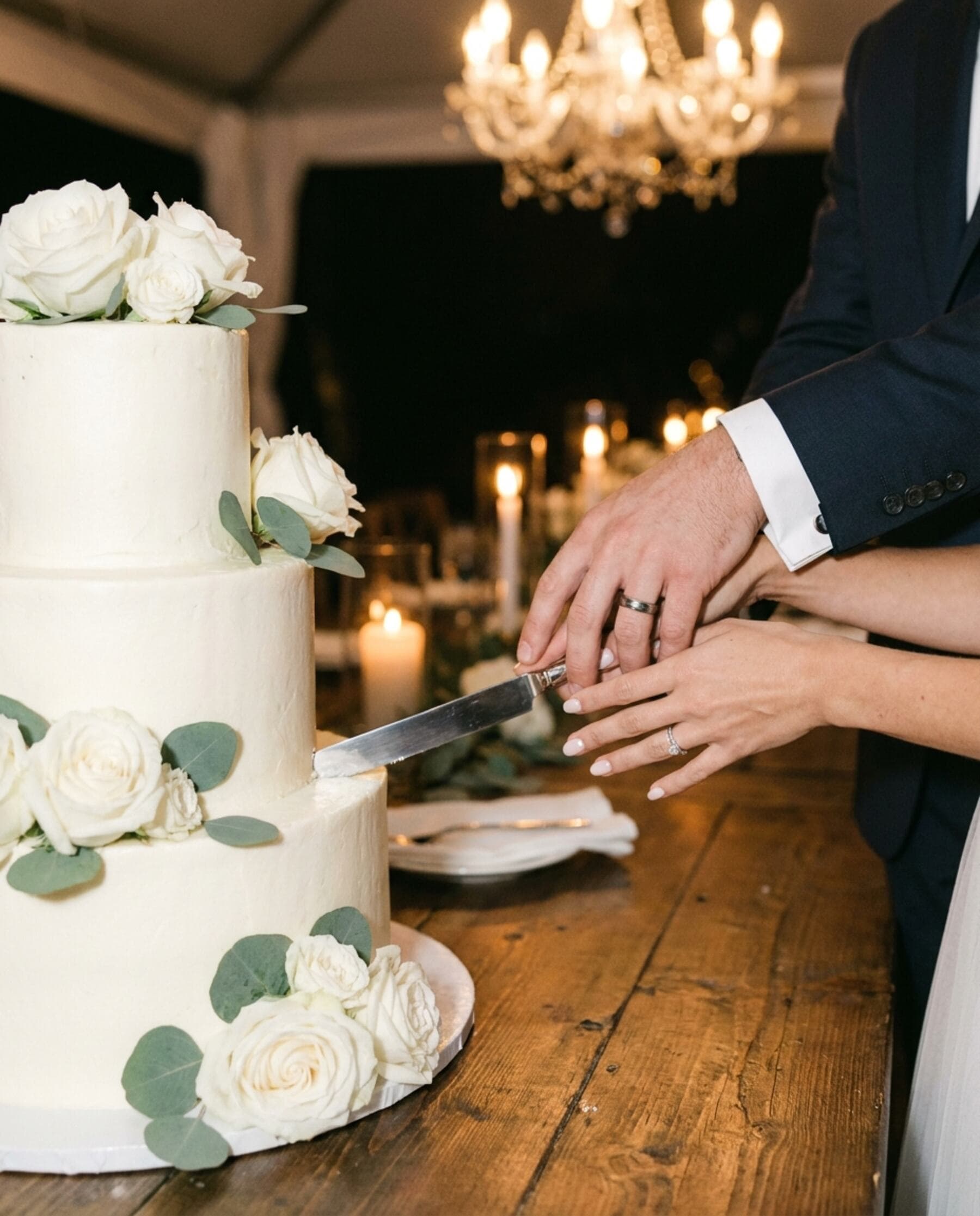 Eine dreistöckige weiße Hochzeitstorte mit Rosen und Eukalyptus, das Brautpaar führt gemeinsam das Messer in die unterste Etage, zu sehen sind nur die Hände