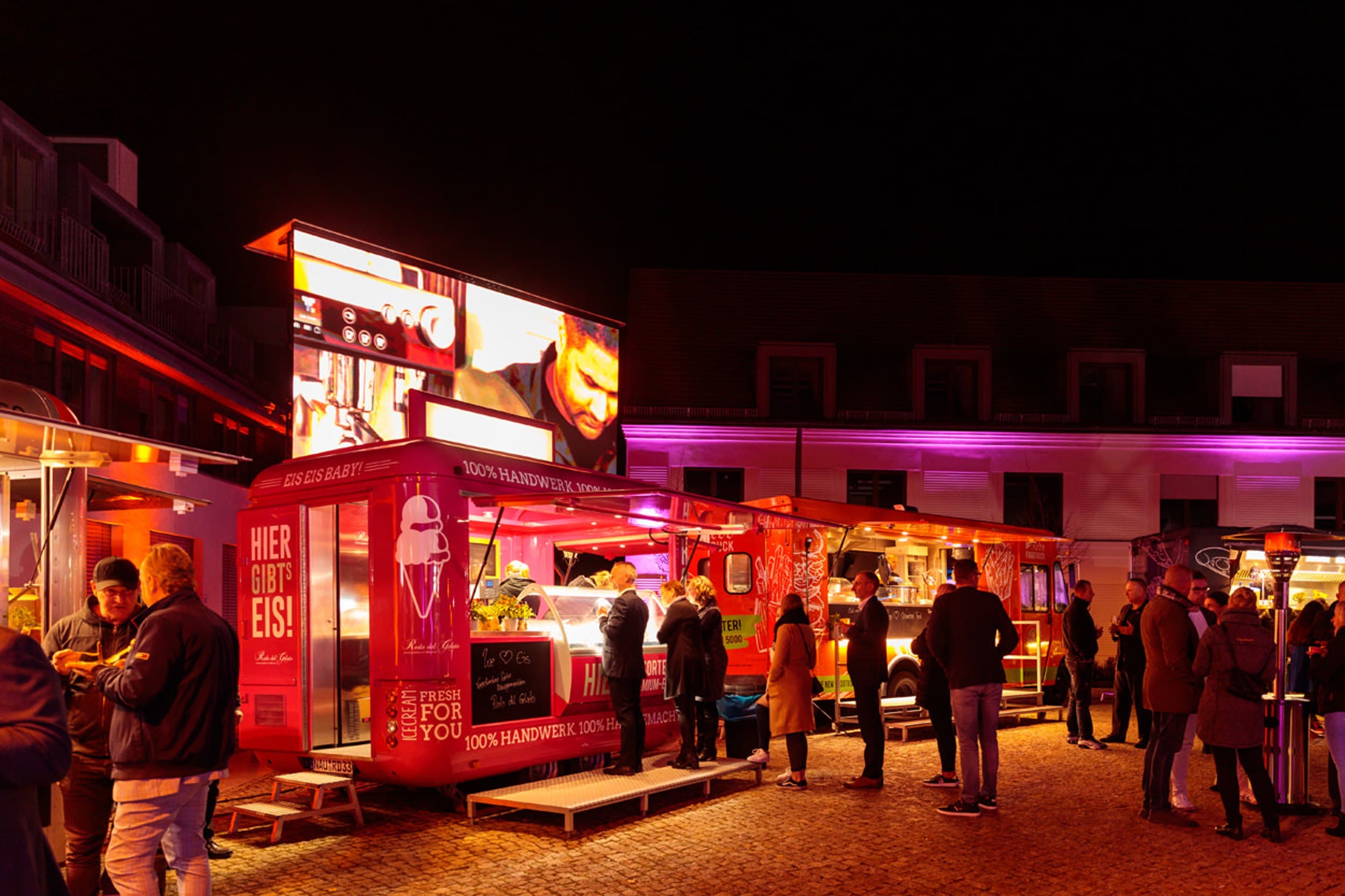 Zwei rot beleuchtete Foodtrucks nebeneinander auf dem Hof bei Nacht, davor wartende Gäste