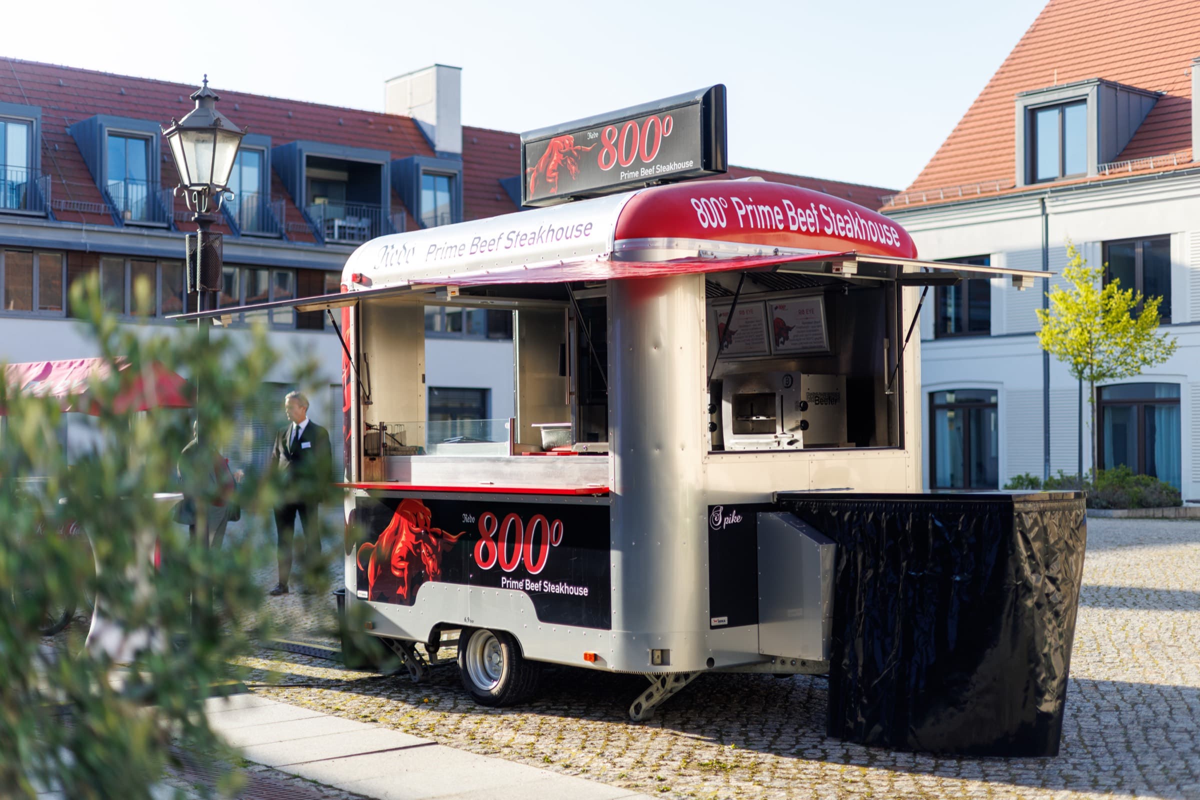Der silberne Foodtruck „800° Prime Beef Steakhouse“ mit rotem Dach auf dem gepflasterten Hof, die Verkaufsklappe offen, dahinter das Gebäude am Neuen Markt