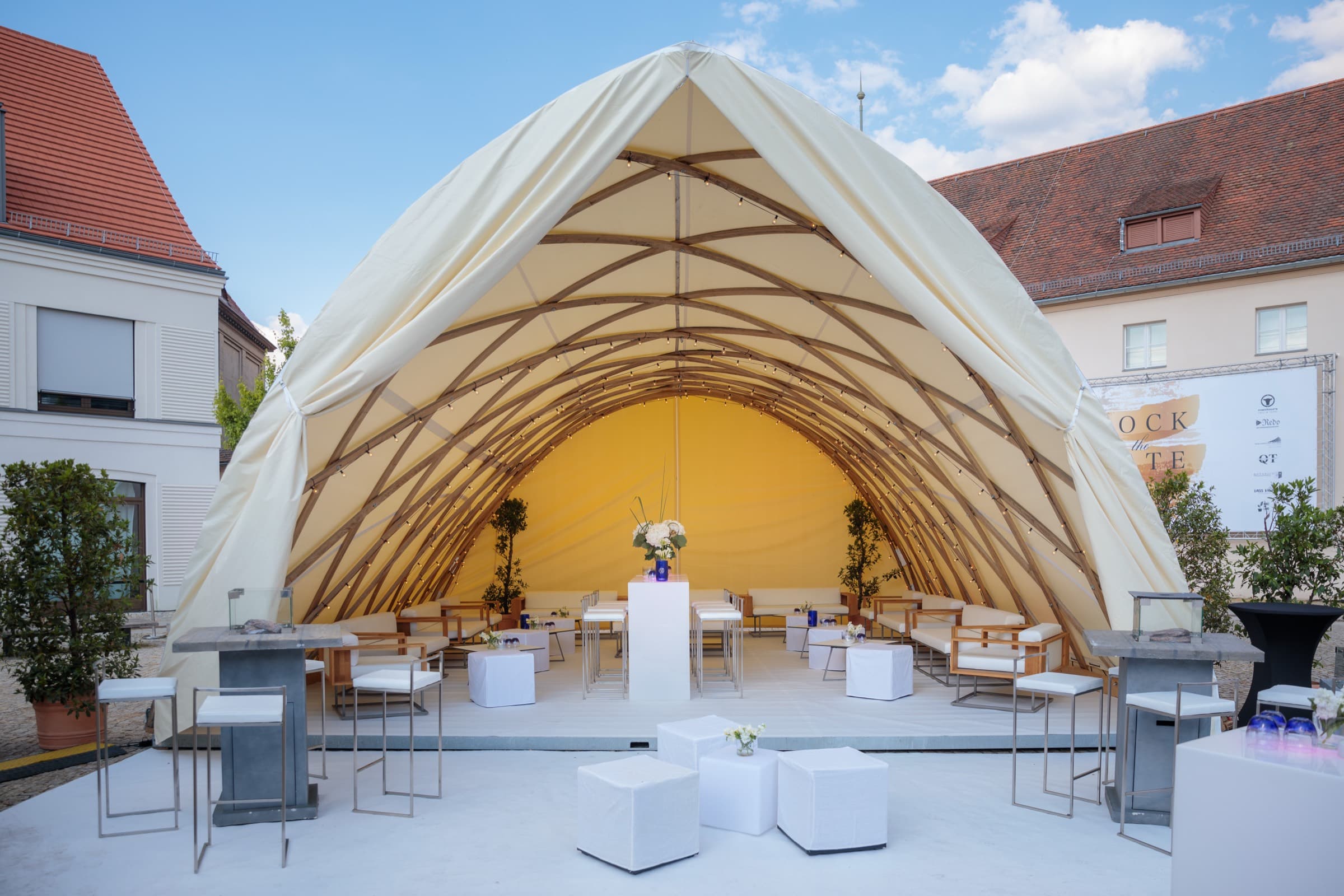 Ein großes Bogenzelt aus hellem Segeltuch über geschwungenen Holzbögen im Hof, darunter eine Lounge mit weißen Sofas, davor Stehtische und weiße Sitzwürfel