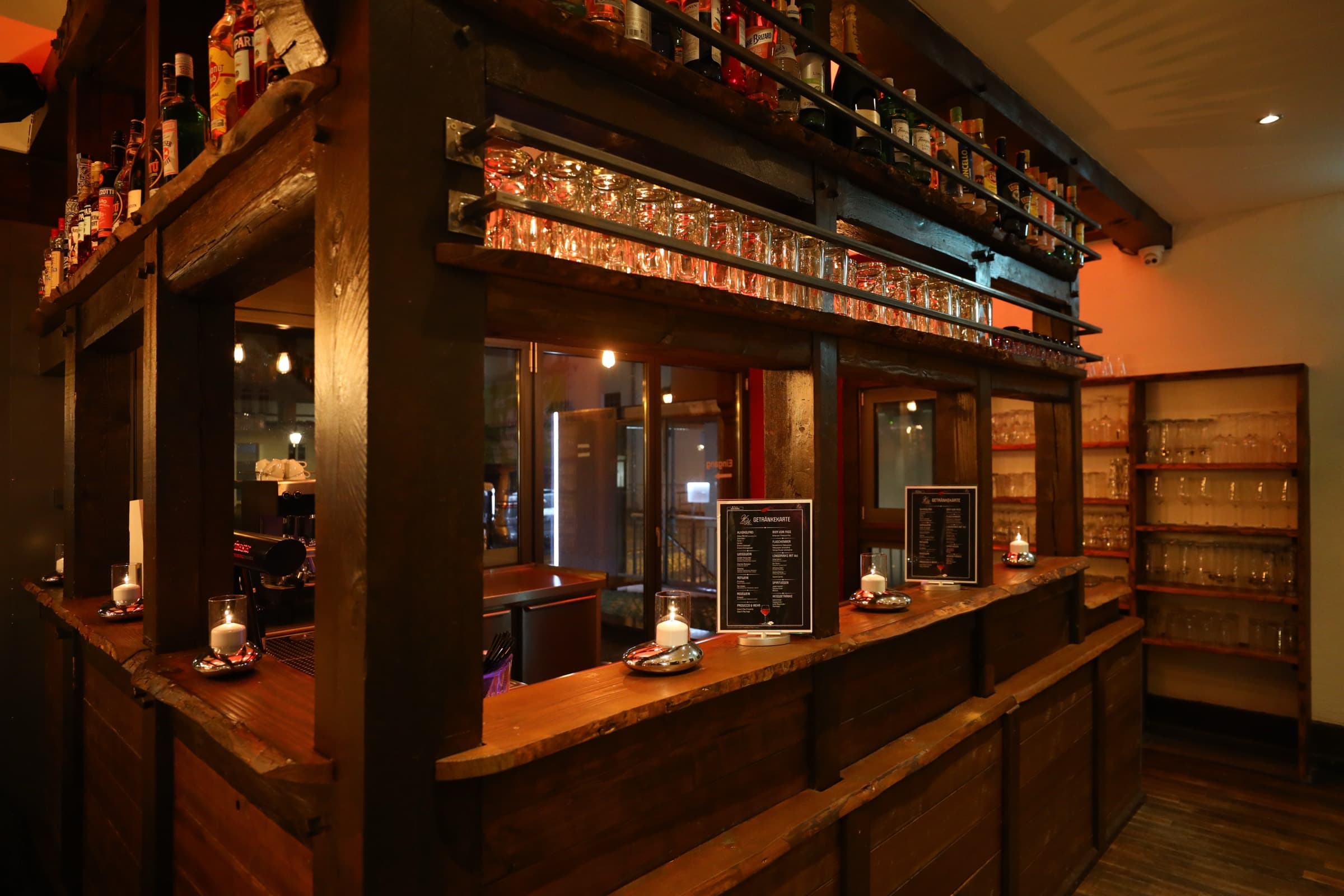 Die Bar aus dunklem Altholz, darüber Reihen von Flaschen und Gläsern im warmen Licht, auf dem Tresen Kerzen und zwei Getränkekarten