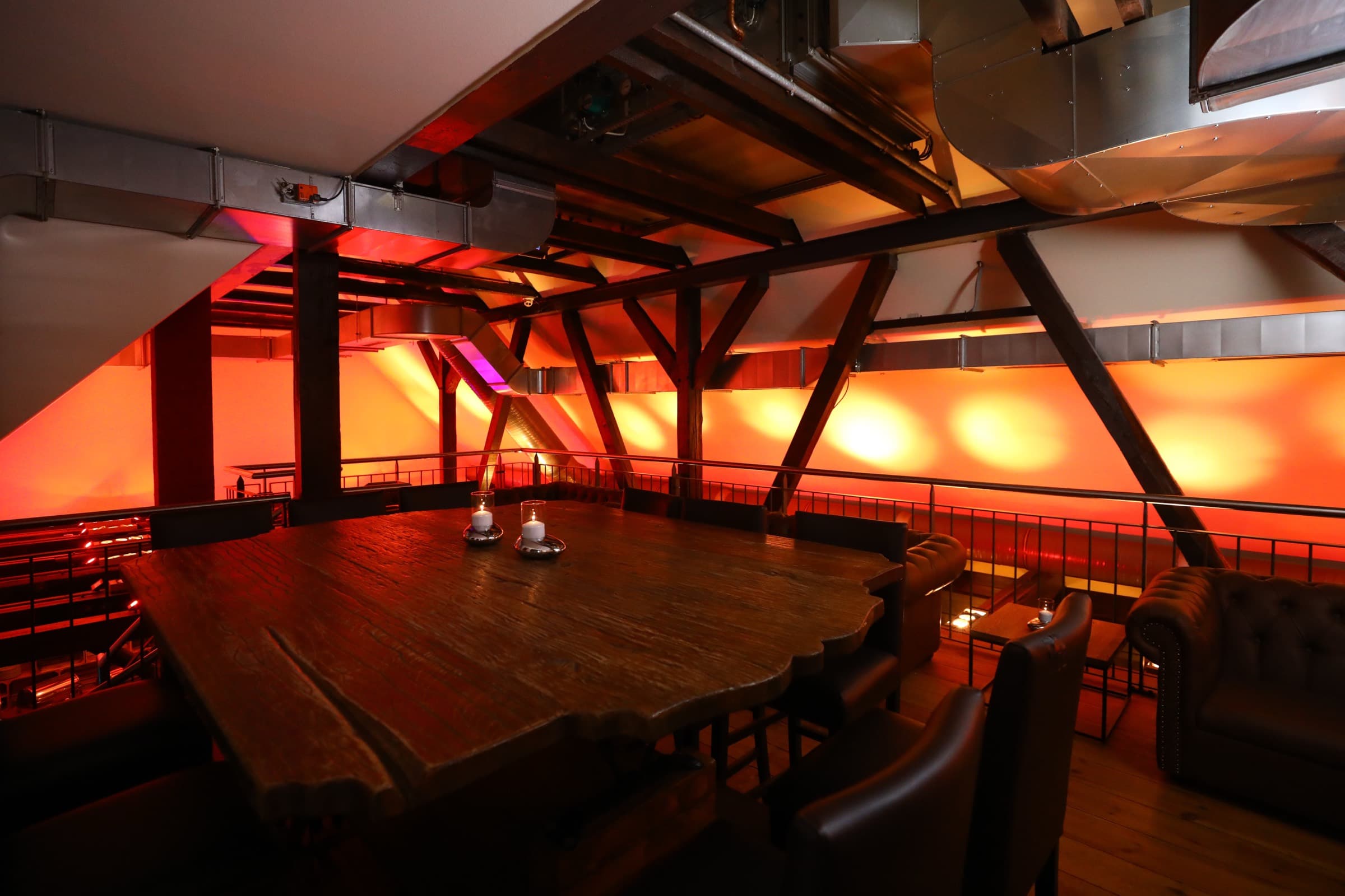 Massiver Holztisch mit Kerzen im Obergeschoss, Ledersessel und eine Chesterfield-Couch an der Galeriebrüstung, offenes Gebälk in Orange angestrahlt