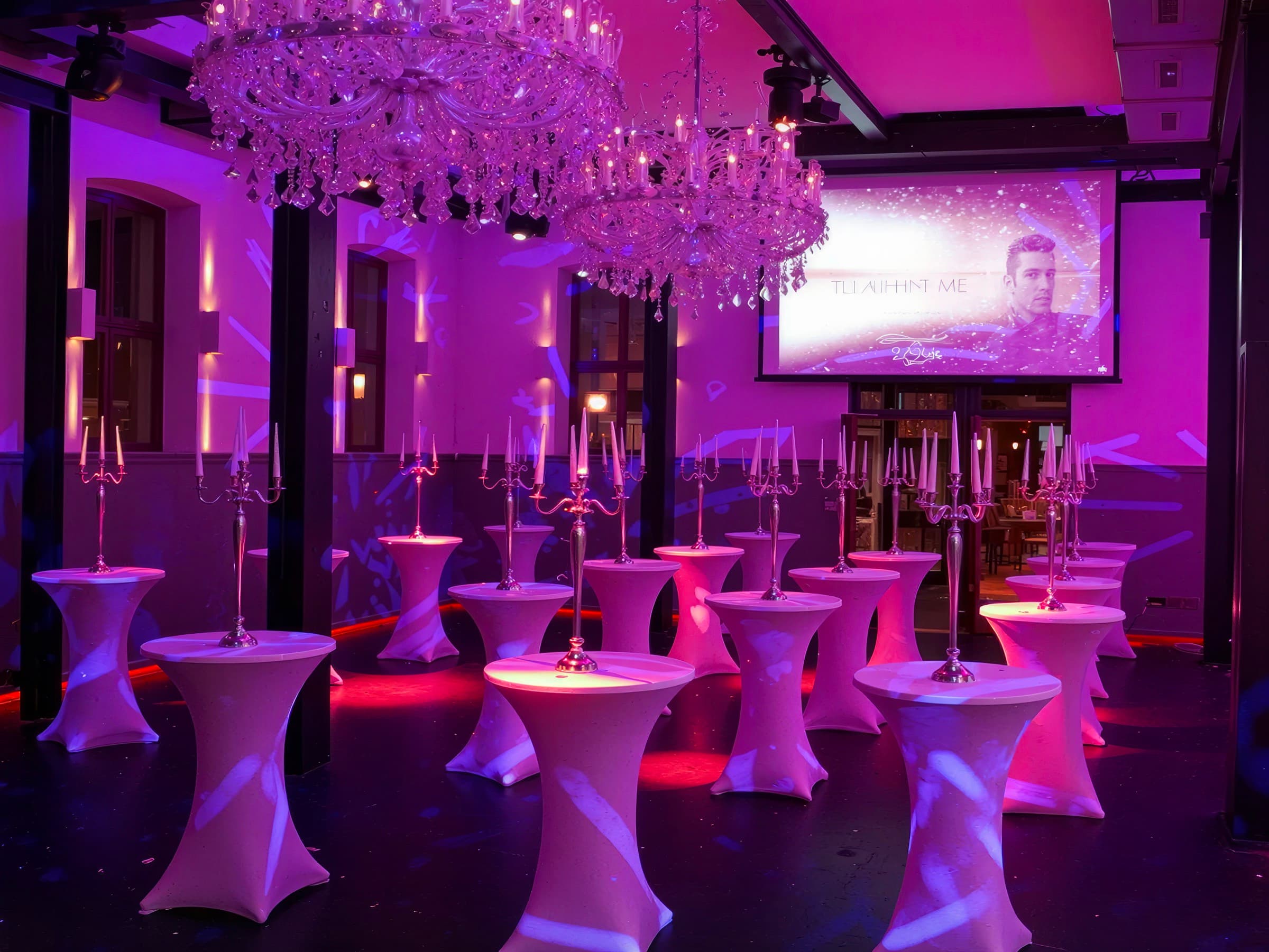 Stehtische mit weißen Hussen und Kerzenleuchtern in Reihen im Saal, im Hintergrund eine Leinwand, der Raum in Violett angestrahlt