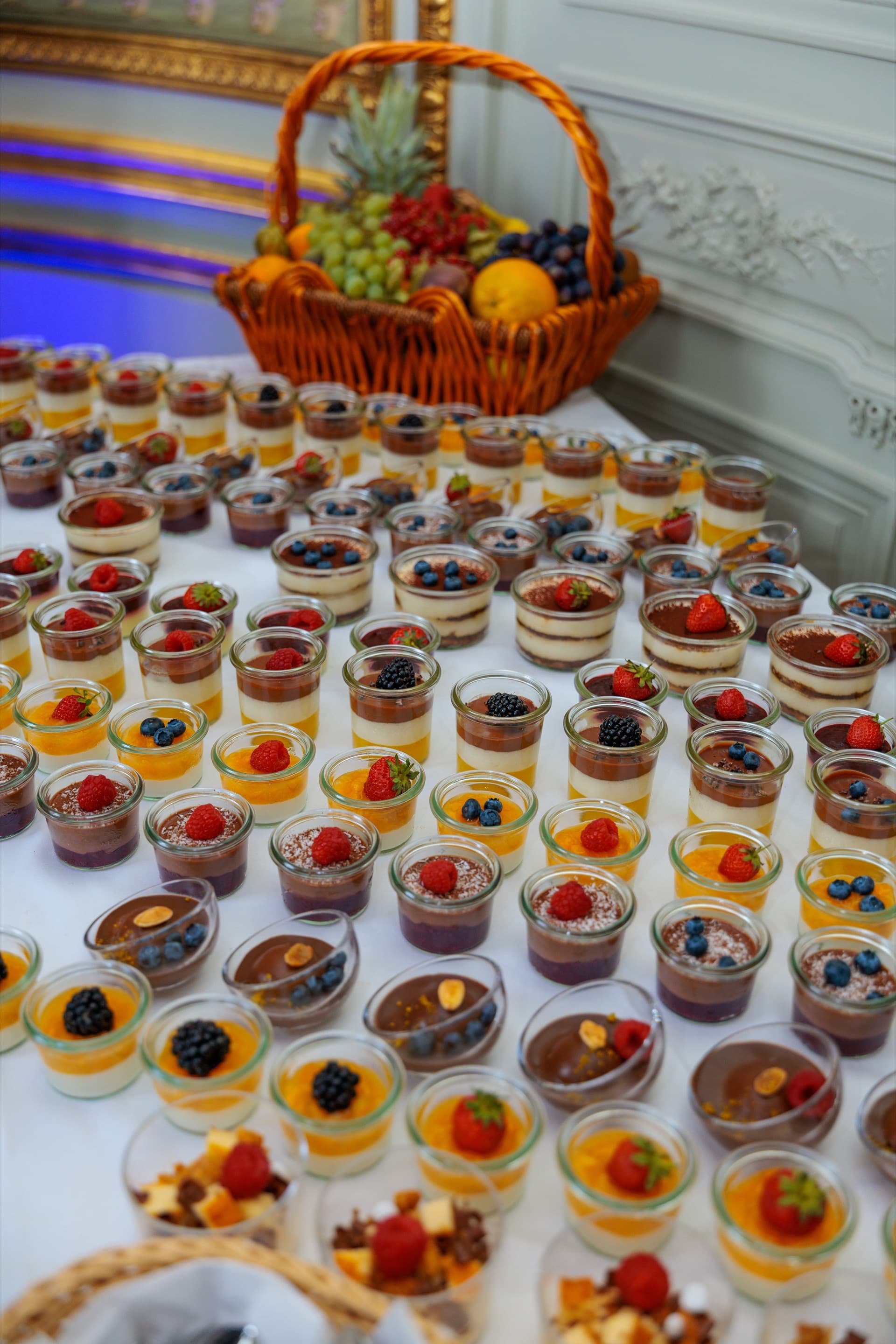 Dessertbuffet aus vielen kleinen Gläsern mit Panna Cotta, Tiramisu und Schokoladenmousse, jedes mit frischen Beeren belegt, dahinter ein Obstkorb