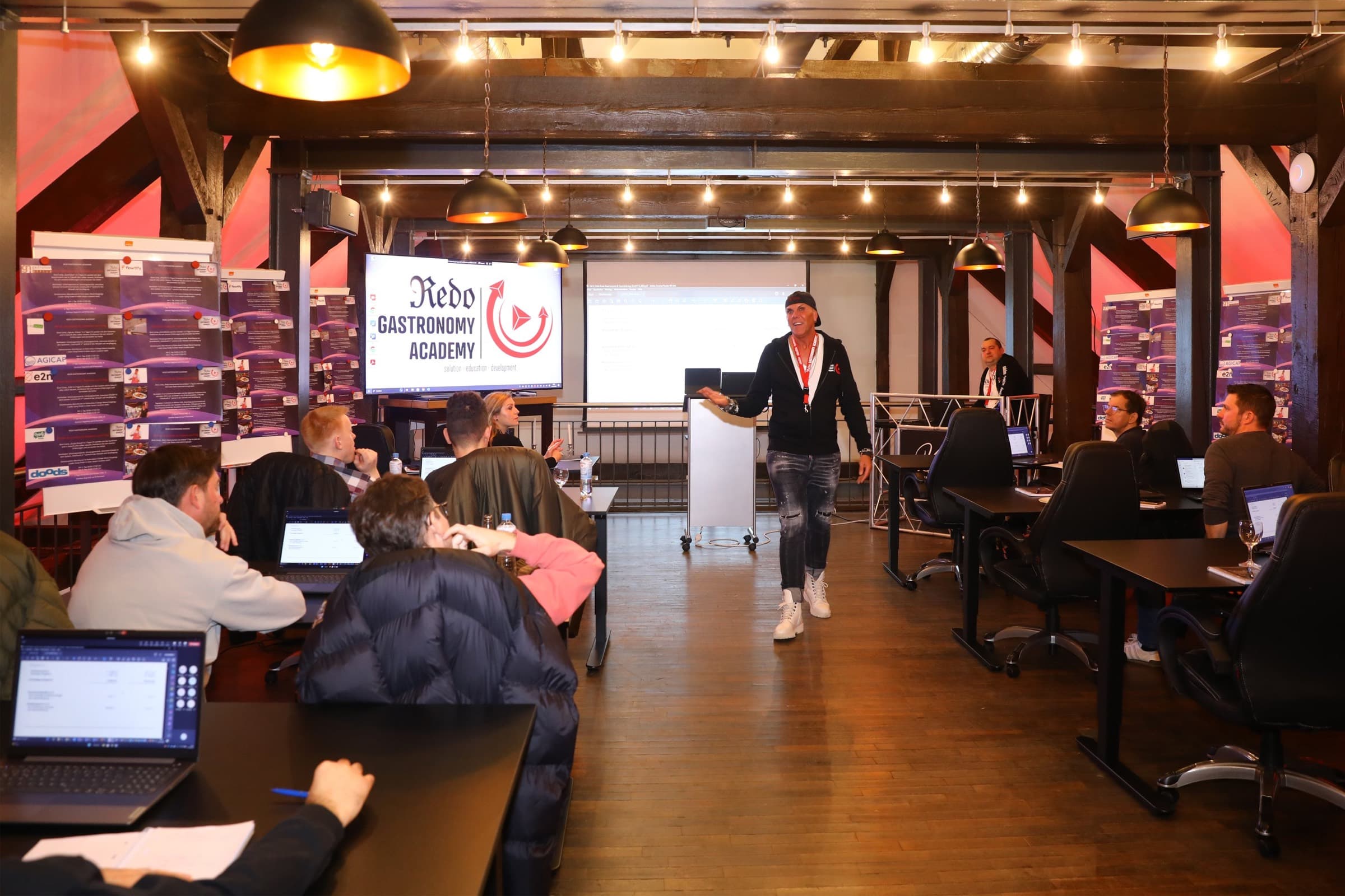 Seminar unter offenem Holzgebälk, Teilnehmer an Tischen mit aufgeklappten Laptops, vorne ein Referent am Stehpult vor der Leinwand der Redo Gastronomy Academy
