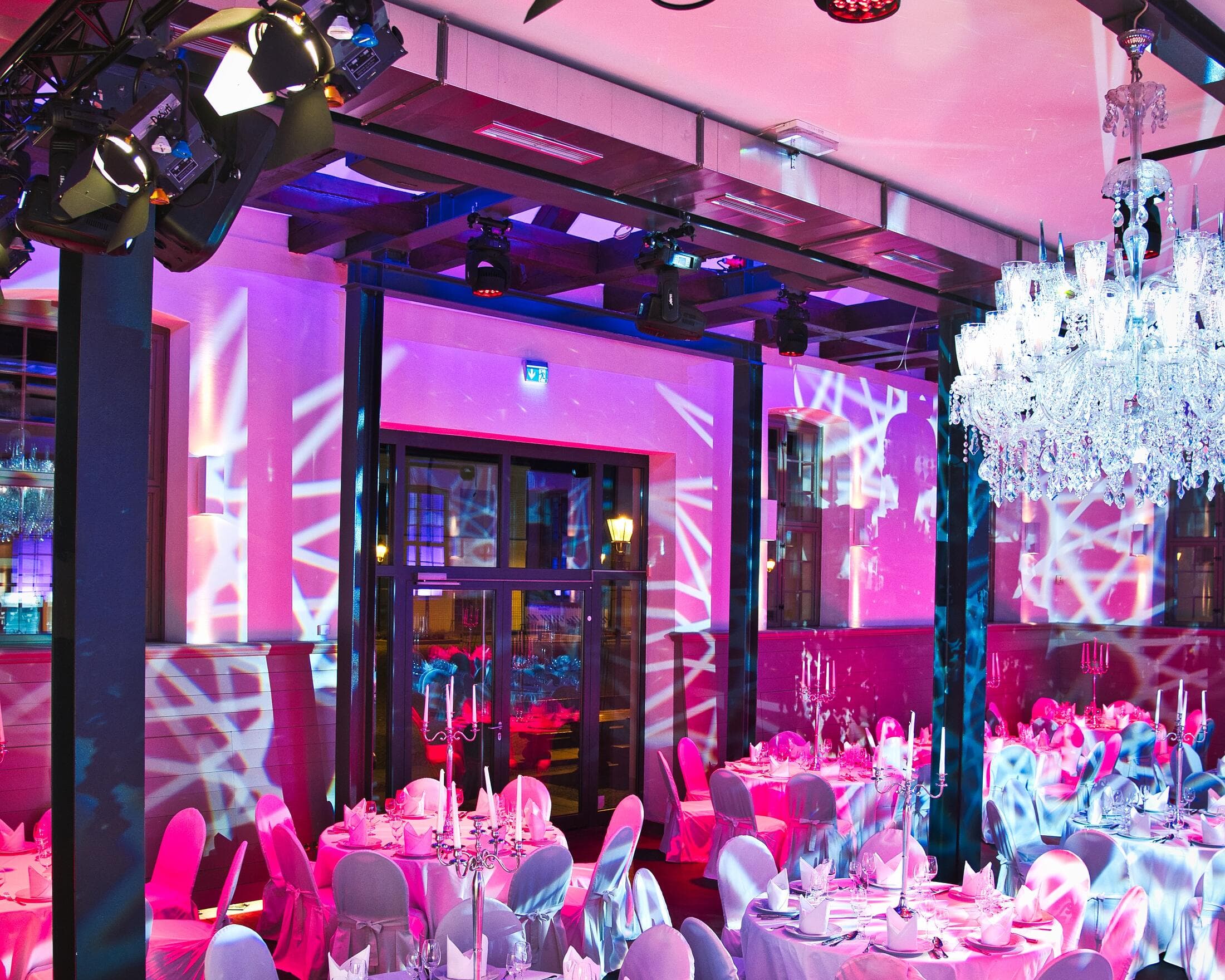 Der Saal in Pink und Violett ausgeleuchtet, oben Scheinwerfer an der Traverse, rechts ein Kronleuchter, an den Wänden weiße Lichtmuster, unten runde Tische mit Stuhlhussen
